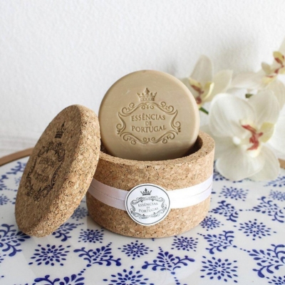 Sabonete redondo beige em embalagem de cortiça com etiqueta branca e flores brancas ao fundo