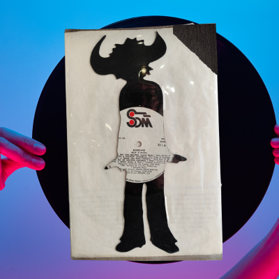 Disco de vinil com capa branca e recorte de figura com chifres, segurado sobre fundo azul