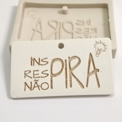 Placa retangular branca em cerâmica com texto gravado e desenho de lâmpada, sobre molde correspondente.
