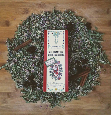 Caixa de chá 'ALL I WANT FOR CHRISTMAS IS TEA' rodeada por folhas secas e paus de canela sobre superfície de madeira.