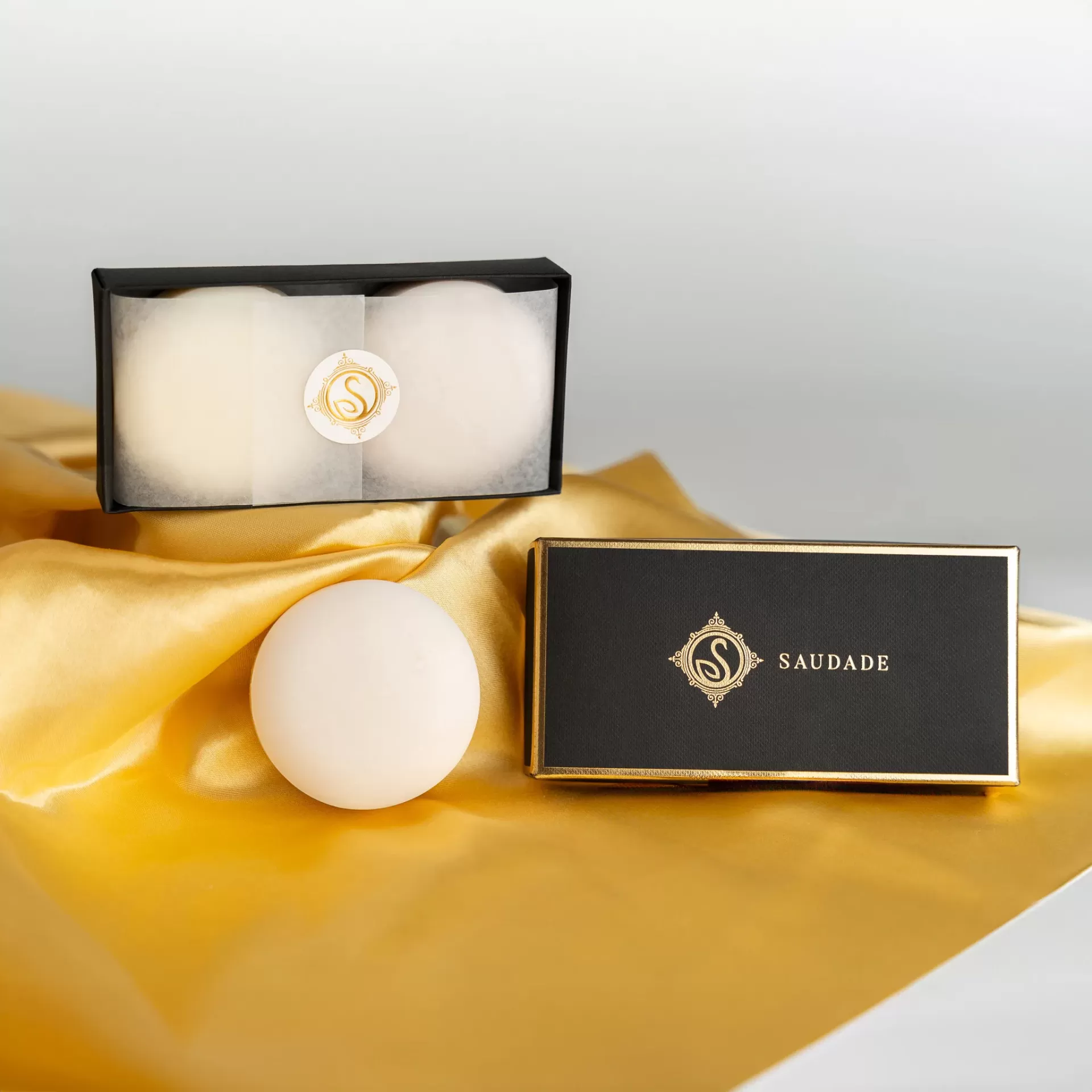 Conjunto de sabonetes brancos em caixa preta com texto SAUDADE sobre tecido amarelo.