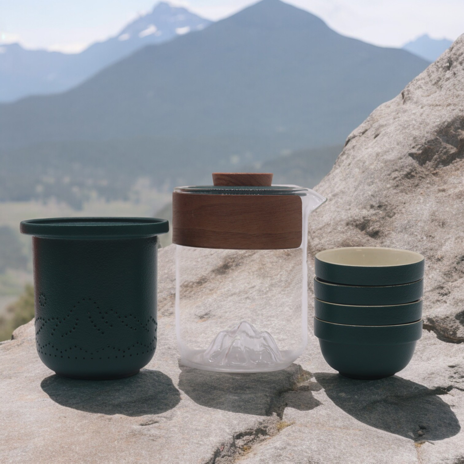 Conjunto de chá ou café com jarra de vidro e tampa de madeira, tigelas verdes empilhadas, sobre pedra com montanhas ao fundo.