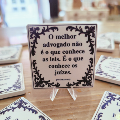 Azulejo quadrado em cerâmica com moldura preta e texto em português sobre advogados, em suporte transparente sobre mesa de madeira