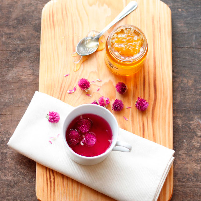 Chávena branca com bebida rosa e flores roxas, frasco de compota alaranjada, colher e pano branco sobre tábua de madeira.