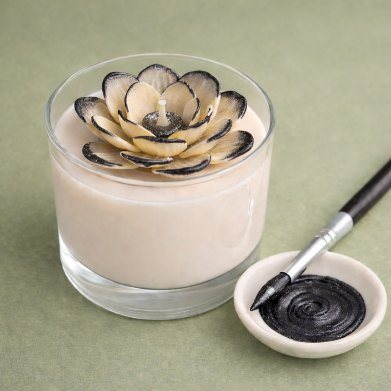 Vela decorativa com flor preta e creme, pincel e tinta preta numa tigela pequena