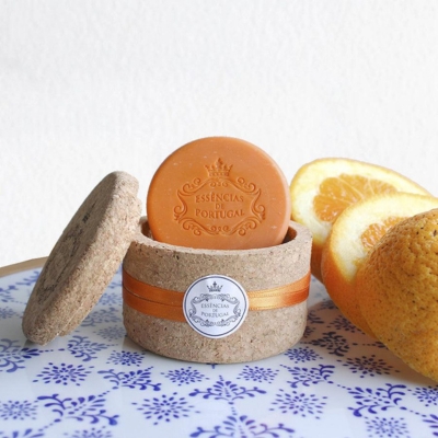 Sabonete laranja em embalagem de cortiça com fita laranja e rótulo branco, junto a uma laranja cortada