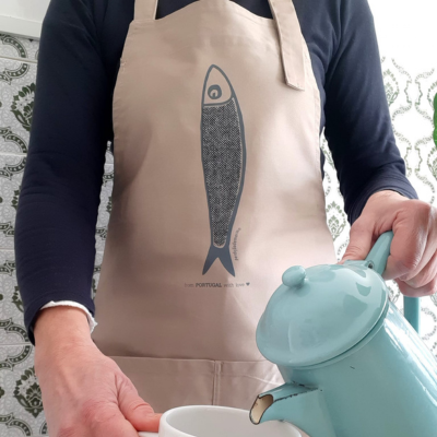 Pessoa com avental bege com desenho de peixe azul a servir chá de bule azul claro para chávena branca