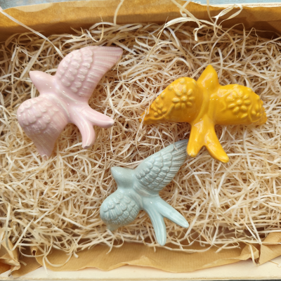 Três pássaros decorativos em cerâmica rosa, amarelo e azul em caixa com palha