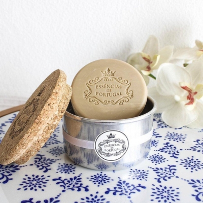 Sabonete redondo cor bege com inscrição, em recipiente metálico com tampa de cortiça, em prato com padrão azul e flores ao lado.