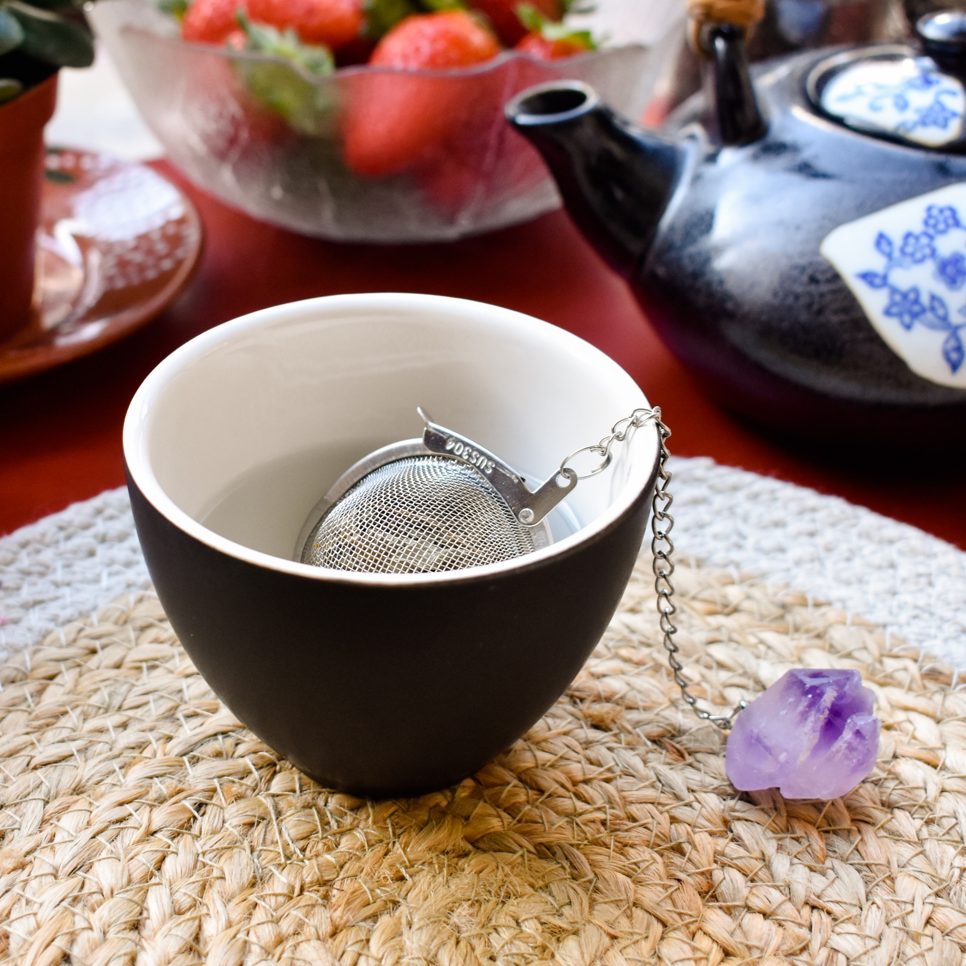 Chávena bicolor com infusor metálico sobre suporte de palha, bule de chá preto com detalhes azuis e tigela com morangos ao fundo.