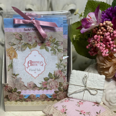 Sache perfumado floral com embalagem transparente, sabonete branco e almofada rosa com flores ao lado