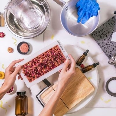 Barra de sabão artesanal com pétalas secas num molde branco rodeada por utensílios de preparação