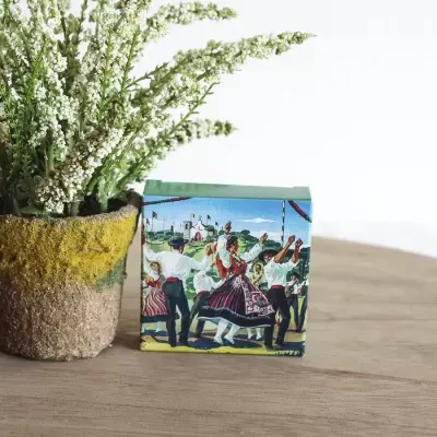 Caixa decorativa com ilustração de dança tradicional portuguesa junto a vaso com flores brancas em mesa de madeira.
