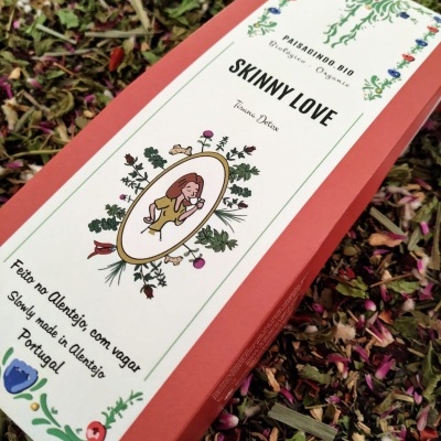 Embalagem vermelha de tisana com rótulo branco decorado e texto em português, rodeada por ervas secas e pétalas.