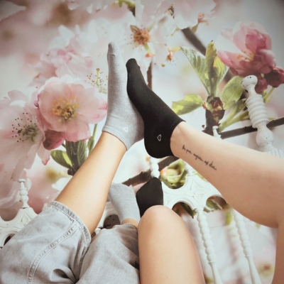 Pares de meias pretas e cinza com corações brancos e pernas levantadas sobre cama com fundo floral.