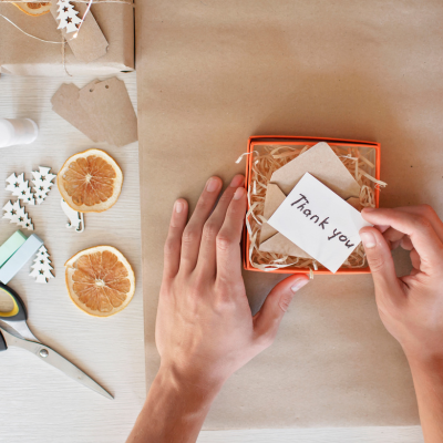 Mãos seguram cartão 'Thank you' sobre caixa laranja com enchimento, ferramentas e decorações para embrulho
