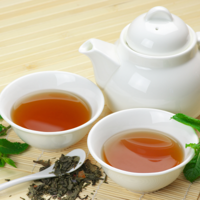 Conjunto de chá branco com chá âmbar, bule branco, folhas de chá soltas e hortelã sobre bambu.