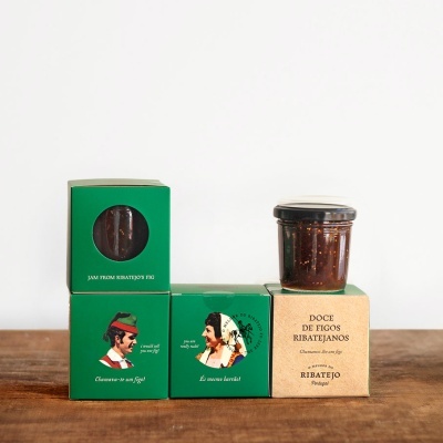 Embalagens de doce de figos com ilustrações e textos em caixas verdes e bege, incluindo frasco de vidro sobre uma mesa de madeira.