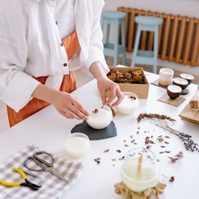 Pessoa a fazer velas artesanais brancas com flores secas numa mesa branca com ferramentas e velas acesas
