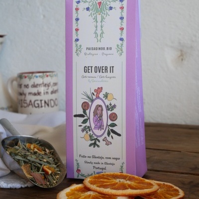 Embalagem de chá orgânico rosa e branco com fatias de laranja desidratada numa mesa de madeira