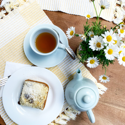 Mesa de madeira com chávena de chá, fatia de bolo e vaso de margaridas.