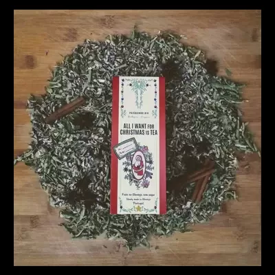 Caixa de chá 'ALL I WANT FOR CHRISTMAS IS TEA' rodeada por folhas secas e paus de canela sobre superfície de madeira.