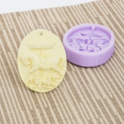 Vela oval amarela com relevo de peixe com coroa ao lado de molde de silicone lilás em tecido riscado bege.