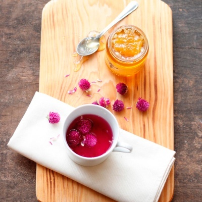 Chávena branca com bebida rosa e flores roxas, frasco de compota alaranjada, colher e pano branco sobre tábua de madeira.