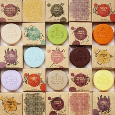 Sabonetes coloridos redondos embalados em caixas de papel com padrões florais e texto em português.