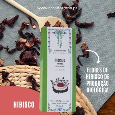 Caixa verde de chá de hibisco com flores secas ao redor sobre mesa com toalha cinza e base de palha