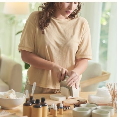 Mulher a fazer sabonetes com t-shirt bege e vários frascos e tigelas sobre mesa de madeira
