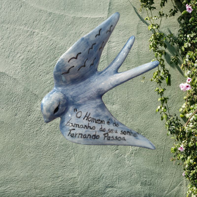 Escultura azul de pássaro em voo com texto de Fernando Pessoa numa parede verde e flores ao lado