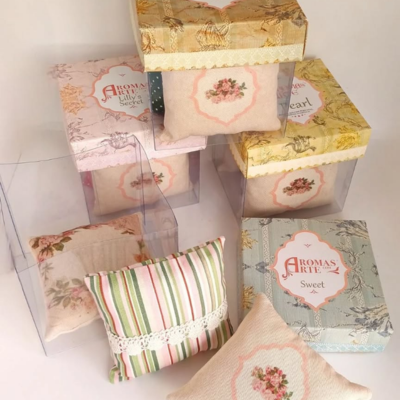 Almofadas decorativas em tecido com padrões florais e riscas, em caixas transparentes com rótulos Aromas Arte.
