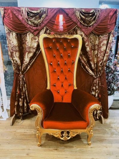 Cadeira clássica vermelha em veludo com estrutura dourada diante de cortinas decorativas castanho e vermelhas