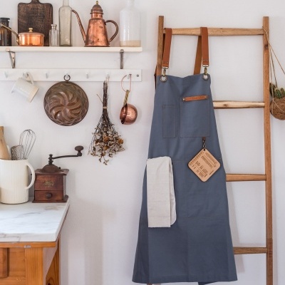 Avental cinzento-azulado com alças de couro pendurado numa escada de madeira junto a utensílios de cozinha
