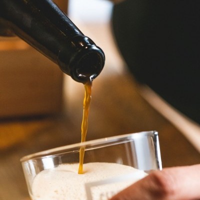 Garrafa preta a verter bebida castanha espumosa para copo de vidro segurado por mão.