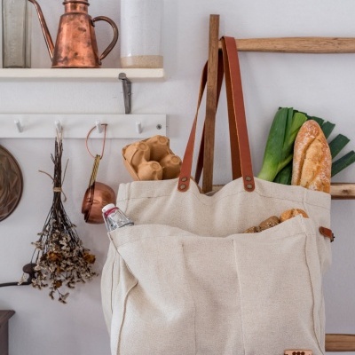 Saco de tecido bege com alças castanhas cheio de pão e legumes pendurado numa escada de madeira, prateleira com jarro cobre e garrafa branca ao fundo