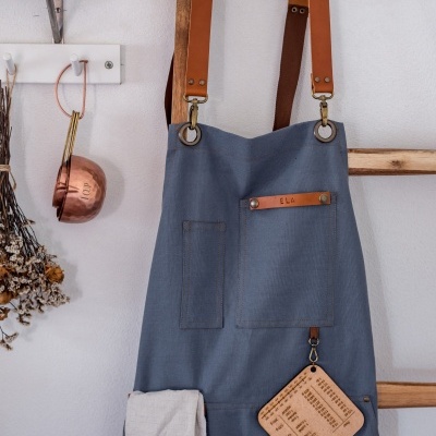 Avental azul de tecido com alças castanhas de couro pendurado numa parede branca com utensílios de cozinha.