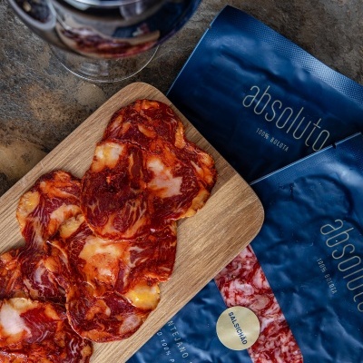 Fatias de salchichón numa tábua de madeira junto a embalagens azuis e copo de bebida escura sob fundo de pedra