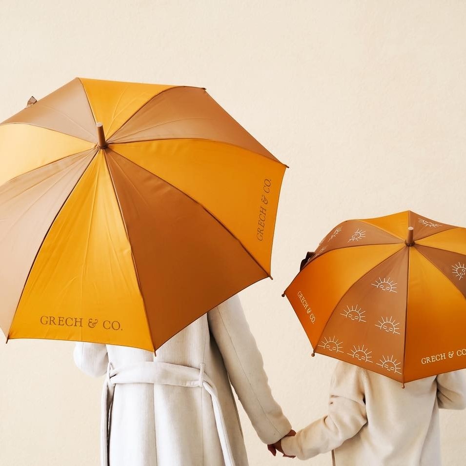 CHAPÉU DE CHUVA SUSTENTÁVEL | MOMMY & ME Dois guarda-chuvas amarelo e castanho com texto GRECH & CO., segurados por pessoas com casacos claros