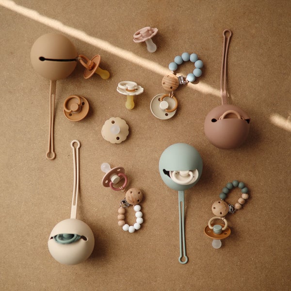 PORTA-CHUCHAS DE SILICONE | MUSHIE Biberões e chupetas em tons pastel com acessórios sobre superfície castanha