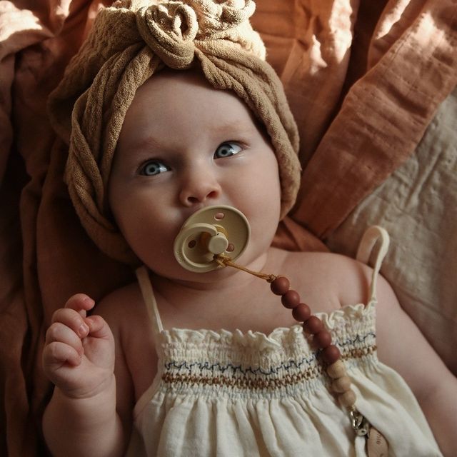 BIBS COLOUR Bebé com chupeta e toucado em tecido castanho claro, vestido branco com bordado, em tecido cor de ferrugem.