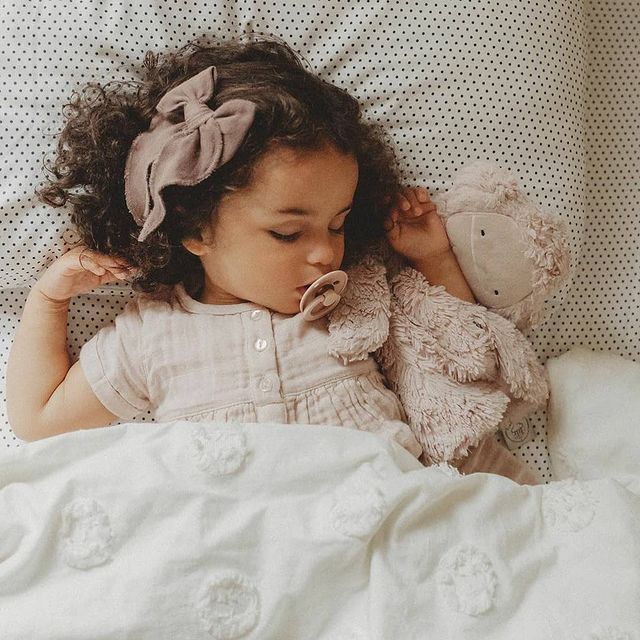 BIBS COLOUR Criança a dormir com chupeta e faixa de cabelo abraçando um boneco cor de rosa