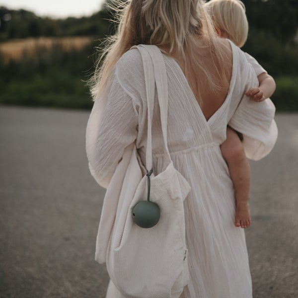 PORTA-CHUCHAS DE SILICONE | MUSHIE Mulher de costas com vestido branco e criança ao ar livre, carregando bolsa com bola verde.
