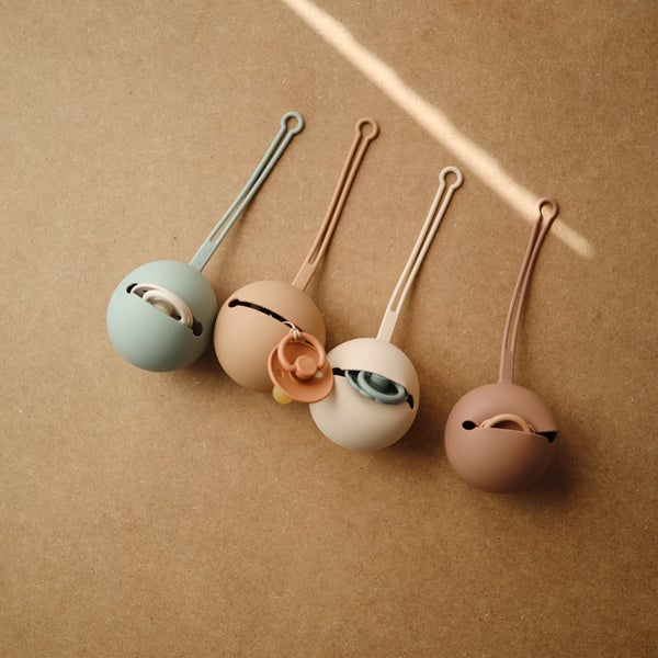 PORTA-CHUCHAS DE SILICONE | MUSHIE Caixas redondas para chupetas em silicone coloridas numa superfície castanha