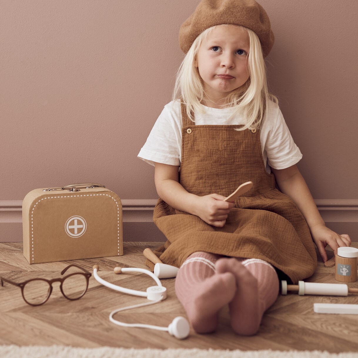 KIT MÉDICO Menina loira com vestido castanho e boina com brinquedos médicos de madeira no chão