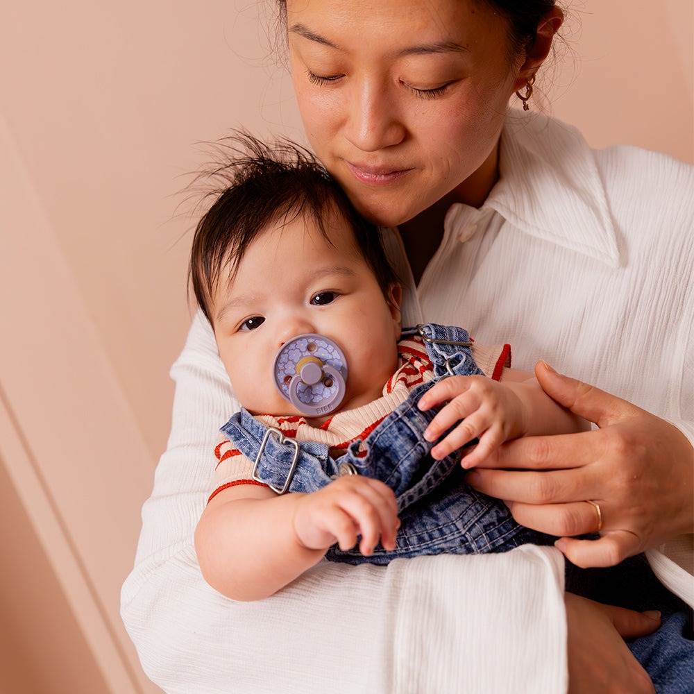 BIBS COLOUR | STUDIO EDITION Mulher com bebé segurando chupeta roxa em ambiente interior