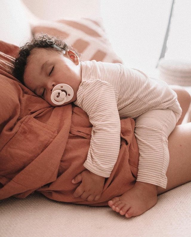 BIBS COLOUR Bebé adormecido com chupeta e pijama listrado a descansar no peito de um adulto.