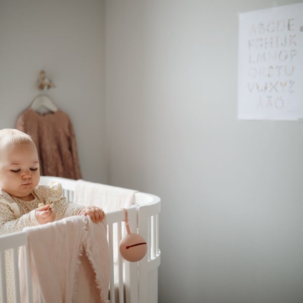 PORTA-CHUCHAS DE SILICONE | MUSHIE Bebé no berço branco com tecido cor de rosa e brinquedo de silicone ao lado, parede cinza com poster de letras.