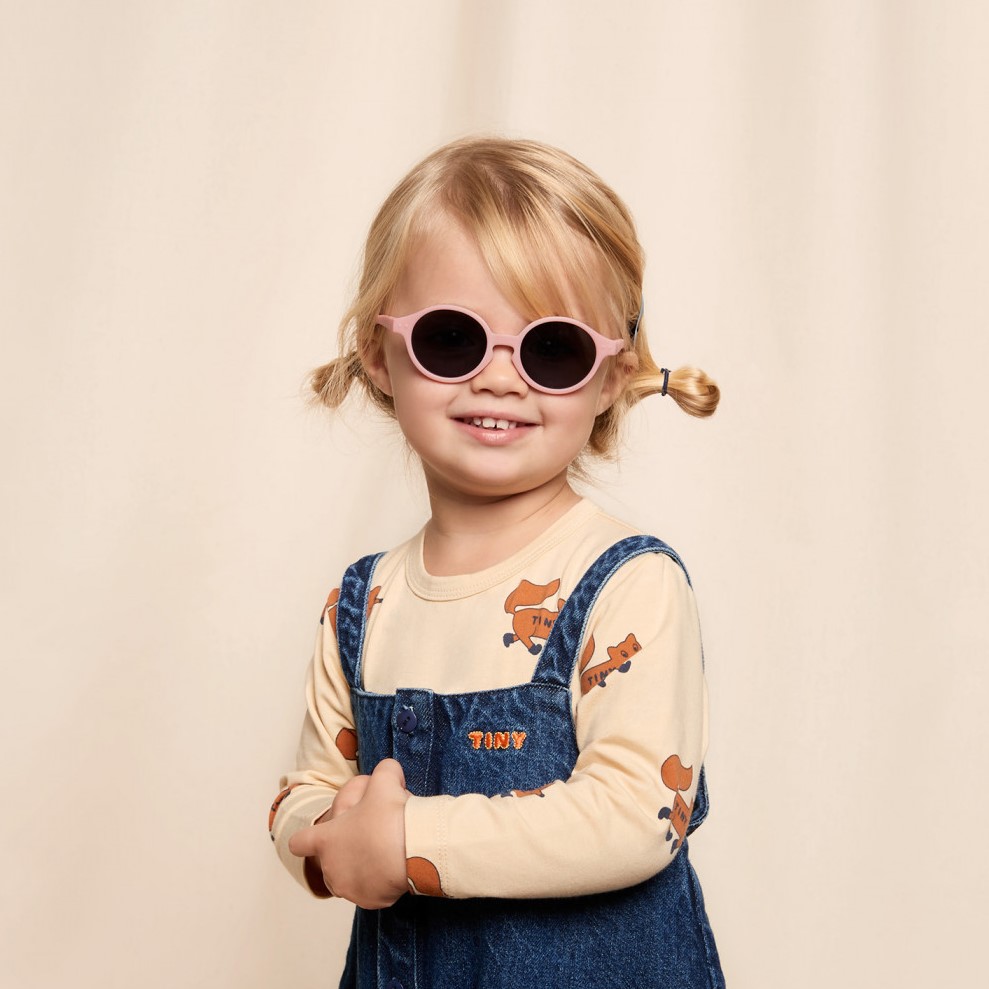 ÓCULOS IZIPIZI | KIDS Criança com óculos de sol rosa e roupa casual em fundo bege